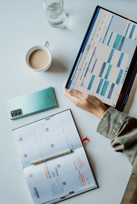 Hands planning dates on a tablet calendar beside a paper planner and coffee